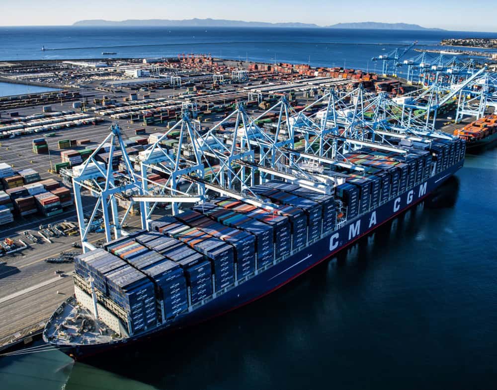 A large CMA CGM container ship docked at a busy port, surrounded by cranes and stacks of shipping containers, highlights global trade efforts much like efforts to take down Lockbit in the digital world, with the ocean and distant land in the background.