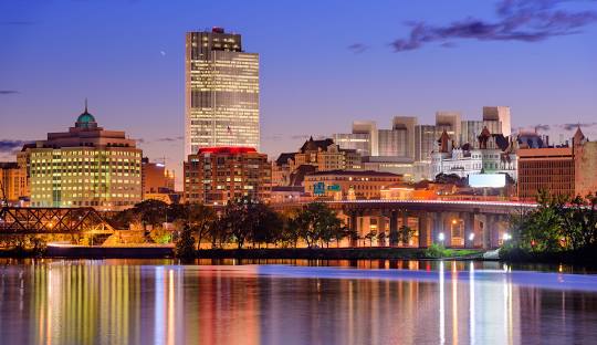 Twilight cityscape with illuminated buildings reflected on a calm river, showcasing the glow of managed IT services in Albany.