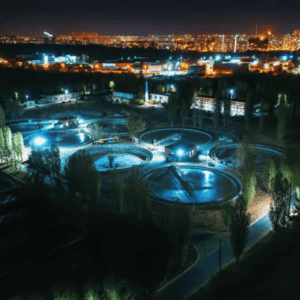 Aerial night view of a wastewater treatment plant with illuminated circular tanks against a cityscape background, emphasizing water security.