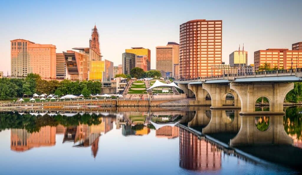 Cityscape at sunrise with modern buildings reflecting in a calm river, a bridge in the foreground managed by IT services Hartford, and clear blue sky.