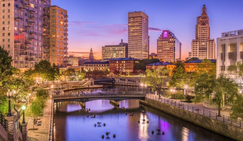 Twilight view over a city's riverwalk with illuminated buildings, a pedestrian bridge, and a tranquil river reflecting city lights near the managed IT services hub in Providence.