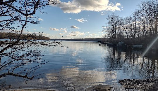 A calm lake surrounded by bare trees on a clear day, with reflections of the sky and clouds on the water's surface, offers a serene escape in Burrillville.