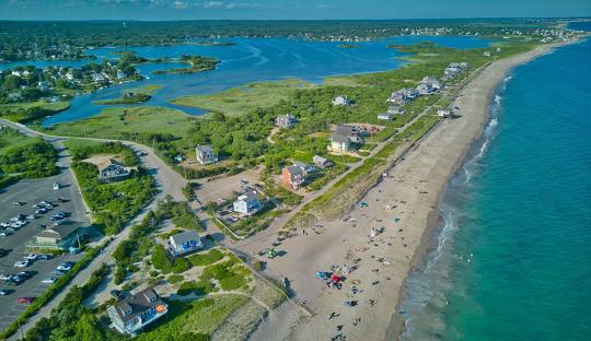 Aerial view of a beach with scattered visitors, houses along the shoreline, a green landscape, and a parking lot on the left, reminiscent of the well-organized approach seen in managed IT services Charlestown provides.