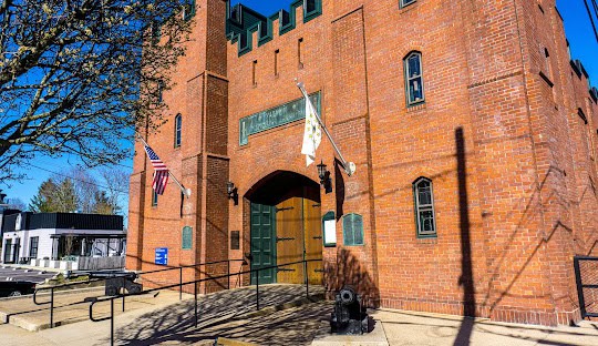 The image shows a red brick building with a castle-like facade, featuring flags on either side of a central door, a ramp leading to the entrance, and a small cannon on the right. This historic site contrasts intriguingly with the modernity of managed IT services in East Greenwich.