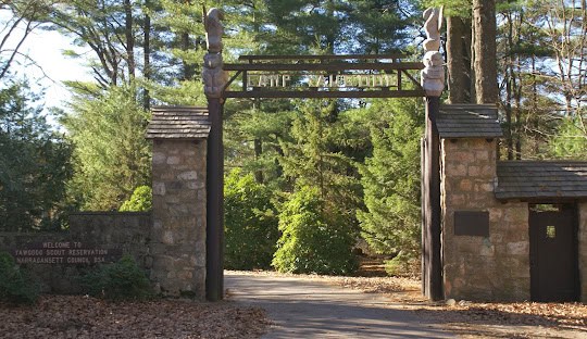 Entrance gate to Yawgoog Scout Reservation, featuring a wooden archway with totem poles on both sides, surrounded by trees and a stone wall. A sign says, "Welcome to Yawgoog Scout Reservation," just a short drive from Hopkinton's managed IT services hub.