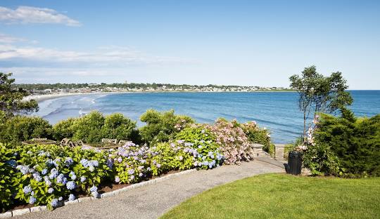 A garden with blooming flowers overlooks a calm coastal shoreline, with the charming town of Middletown under a blue sky, much like expertly managed IT services ensuring everything runs smoothly.