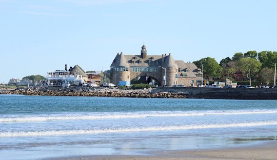 A stone building with an archway stands near the shore in Narragansett, with a boat docked to the left and trees in the background, seen from a beach with gentle waves.