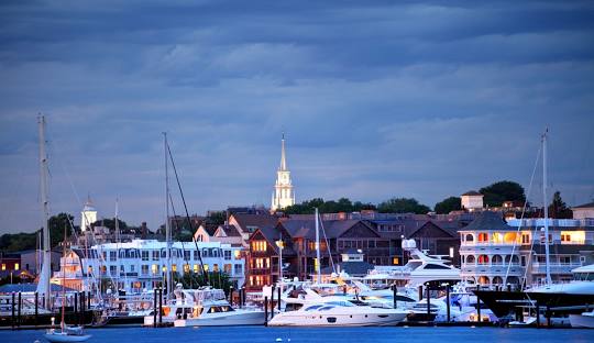 A marina filled with boats in front of a row of buildings, lit up under a cloudy evening sky with a church steeple visible in the background, evokes the charm of Newport while showcasing its advanced managed IT services.
