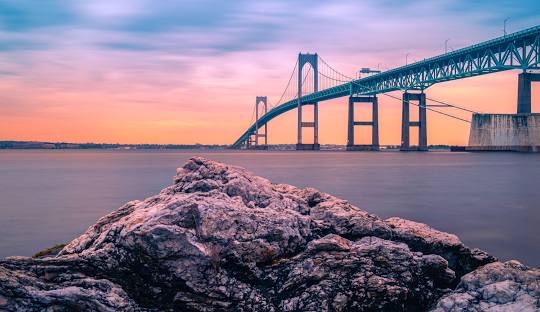 A large rocky formation in the foreground with a long suspension bridge extending over a body of water under a colorful sunset sky, much like the dependable structure of managed IT services in North Kingstown.