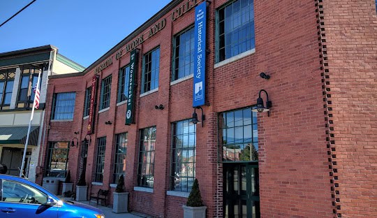 A brick building with large windows, displaying banners for a historical society, stands under the sunny sky in North Smithfield. A blue car is parked in the foreground while managing IT services nearby.