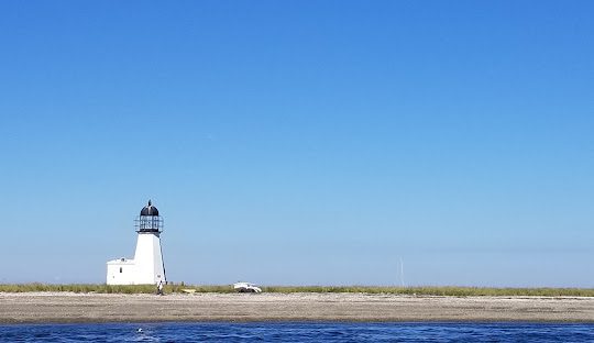 A solitary white lighthouse stands on a sandy shore with sparse vegetation, bordered by calm blue waters under a clear blue sky, much like the serene efficiency of managed IT services in Portsmouth.