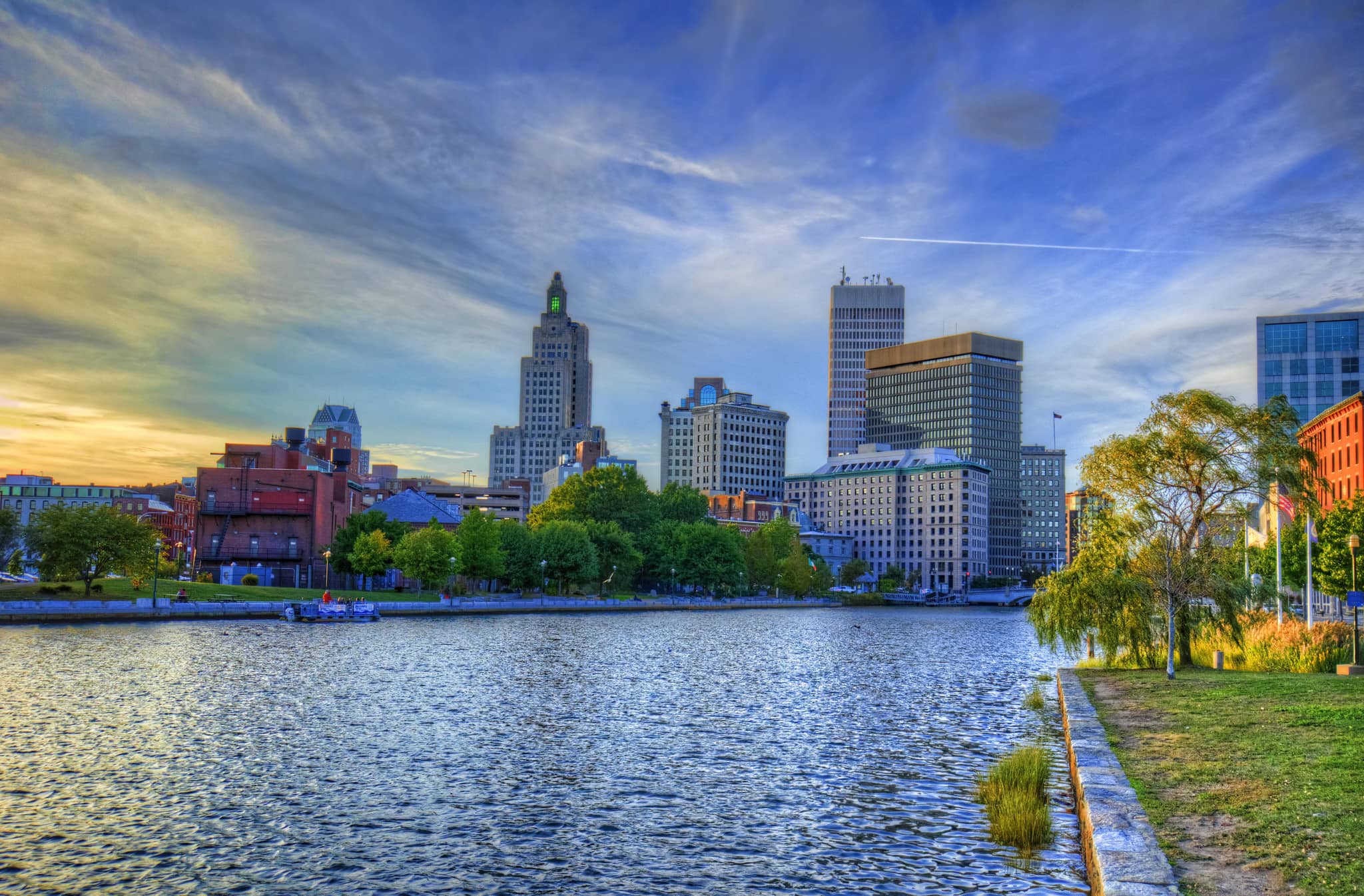 Sunset view of a city skyline with contemporary buildings reflected in a river, under a partly cloudy sky.