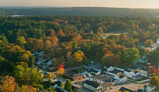 Aerial view of Scituate with residential houses and buildings surrounded by dense trees displaying autumn foliage. A forest and distant hills are visible in the background, hinting at the serene beauty of the town that also offers robust managed IT services.