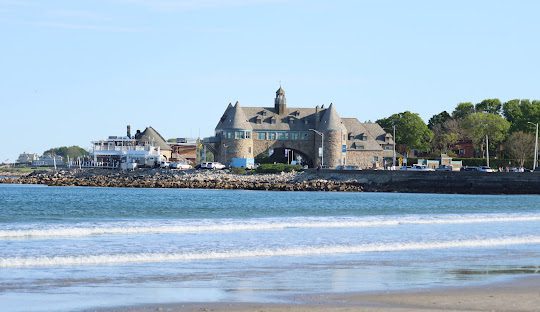 A stone building with a central archway sits along a rocky shoreline in South Kingstown, with a large boat and other buildings in the background, all under a clear sky.