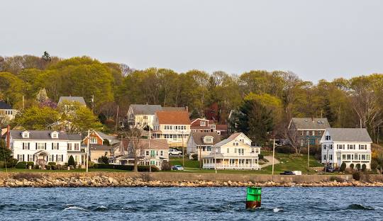A row of houses along a tree-lined shore with a green buoy visible in the foreground on a body of water, reminiscent of the tranquility you’ll find when considering managed IT services in Tiverton.