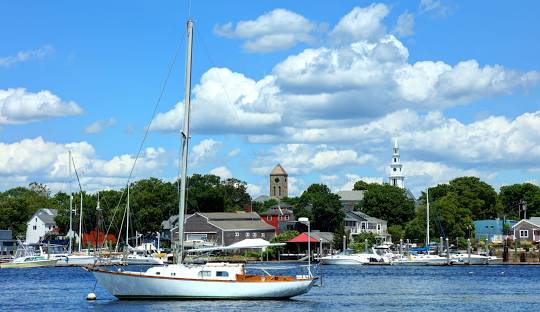 A sailboat is anchored in a calm harbor with various buildings and trees in the background under a partly cloudy sky, much like how managed IT services keep businesses in Warren stable amidst digital turbulence.
