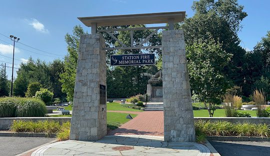 A stone archway entrance to Station Fire Memorial Park, featuring a wooden sign. The park, located near managed IT services in West Warwick, appears well-maintained with greenery, benches, and a pathway leading further into the serene haven.