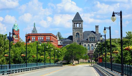 A view of a bridge leading to the historic town of Woonsocket, with red brick and stone buildings, surrounded by green trees and street lamps on a clear day.