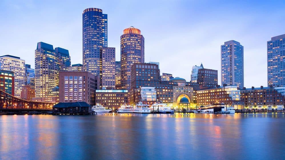 Twilight skyline of Boston with illuminated buildings reflected in the water, featuring a prominent round, brick building and a pedestrian bridge.