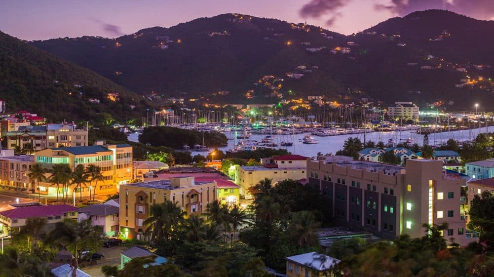 Twilight view of a picturesque coastal town with illuminated buildings and a vibrant harbor full of boats, encircled by lush green mountains.