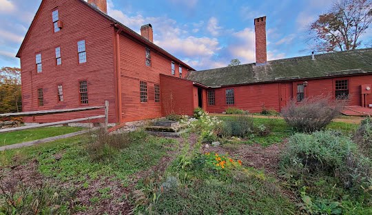 A red wooden house is surrounded by greenery and a garden, with a wooden fence on the left and a chimney protruding from its roof. The sky, over Andover CT, is partly cloudy.