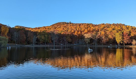 A lake with reflections of autumn trees and a clear blue sky, people in paddle boats, and a hill with a structure in the background—reminiscent of the serene landscapes you could explore while discussing managed IT services in Berlin CT.