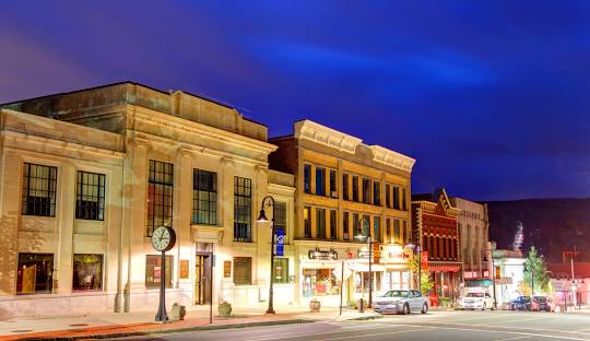 A nighttime view of a small town street in Bristol CT with illuminated historic buildings, storefronts, cars parked along the sidewalk, and street lights casting a warm glow, perfectly showcasing the charm of this town known for its managed IT services.