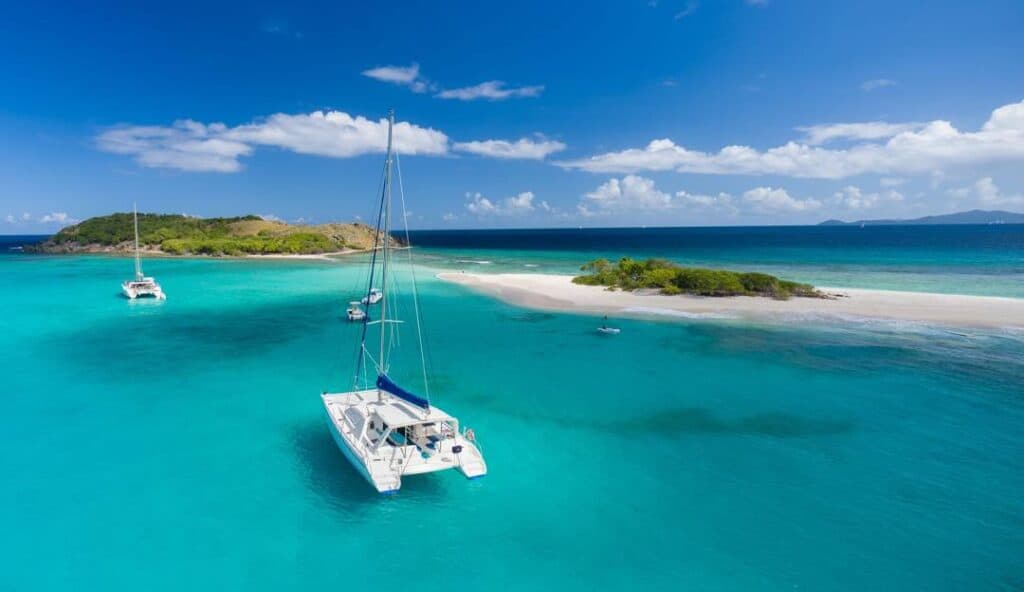 A catamaran is anchored in the clear blue waters of the British Virgin Islands near small islands with white sandy beaches, under a blue sky with scattered clouds.