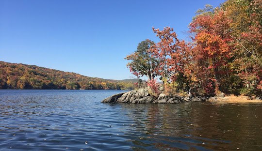 A calm lake with rocky shoreline and colorful autumn trees under a clear blue sky. Hills covered in fall foliage can be seen in the background, resembling the tranquility you'll find when employing managed IT services in Brookfield, CT.