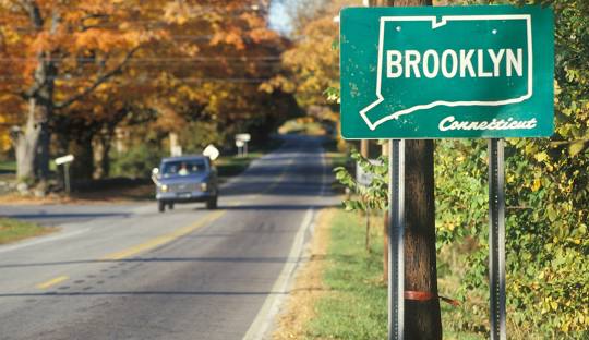 Road sign for Brooklyn, Connecticut, beside a two-lane road with trees in autumn colors and a car driving in the background. Just as serene as our managed IT services in Brooklyn, CT.