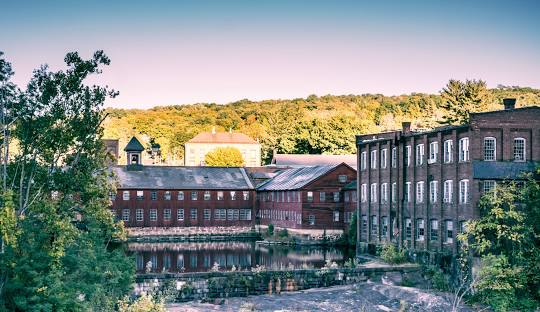 A 19th-century mill complex sits by a calm river in Canton, CT, with red brick buildings surrounded by trees in early autumn, embodying the charm of New England while seamlessly integrating modern managed IT services.