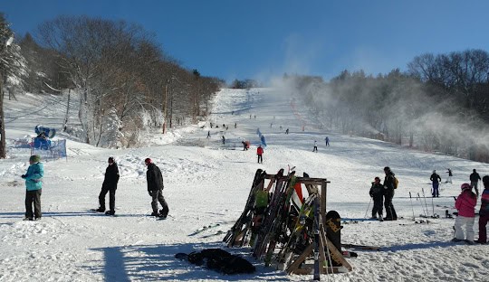 Skiers and snowboarders on a snow-covered ski slope in MA with trees in the background. A rack with skis and poles stands in the foreground, capturing the essence of a perfect winter day.