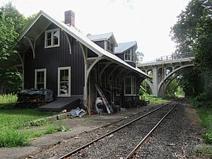 A rustic train station building stands beside railroad tracks, with a tall arched bridge in the background. Piles of wood are stacked near the station, while greenery surrounds the area. In Cornwall CT, even this tranquil scene could benefit from managed IT services to keep things on track.