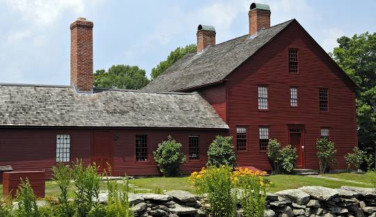 A red-painted, two-story historic house with multiple chimneys and windows stands proudly, surrounded by a well-maintained garden and a stone fence. This charming property in Coventry CT also offers the added convenience of managed IT services.