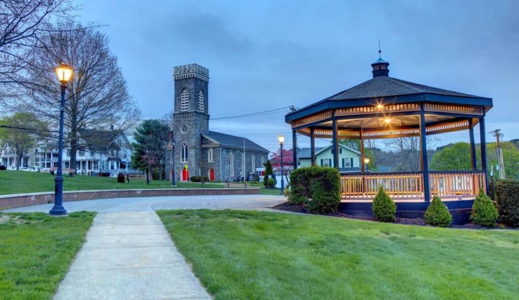 A gazebo in a park with a paved walkway, lamp post, and a church with a bell tower in the background, under a cloudy sky at dusk. The serene setting is near Derby CT, evoking the same sense of reliability one expects from managed IT services.