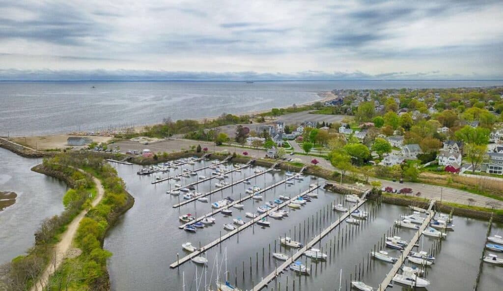 Aerial view of a marina with docked boats, surrounded by green land, residential houses, and a body of water extending towards the horizon under a cloudy sky. Nearby, the efficient tech solutions offered by managed IT services in Fairfield, CT ensure seamless operations for local businesses.