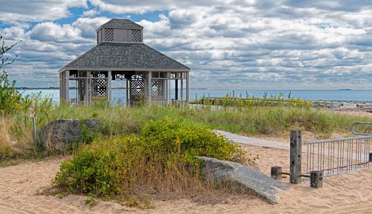 A wooden gazebo stands on a sandy beach with grass and plants in the foreground, under a partly cloudy sky, offering an idyllic setting for companies providing managed IT services in Madison, CT.