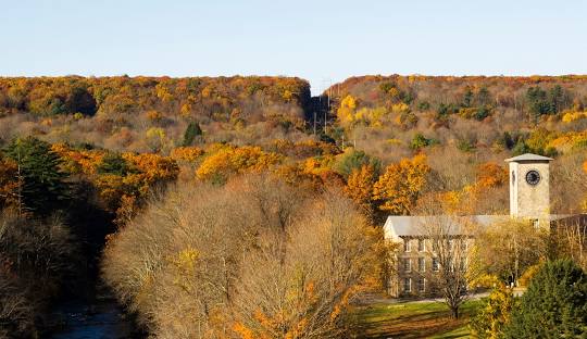 A building with a clock tower stands amidst trees with autumn foliage near a stream, with a hill in the background, reminiscent of the tranquil beauty found in Mansfield CT, home to exceptional managed IT services.