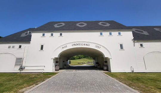 A pale, barn-like building with "Ommegang" and the years 1549 and 1997 written above an arched entrance. The building features a cobblestone path leading through the arch. Under the clear blue sky, it stands as a quaint testament to tradition, much like managed IT services in Maryland ensure modern efficiency.