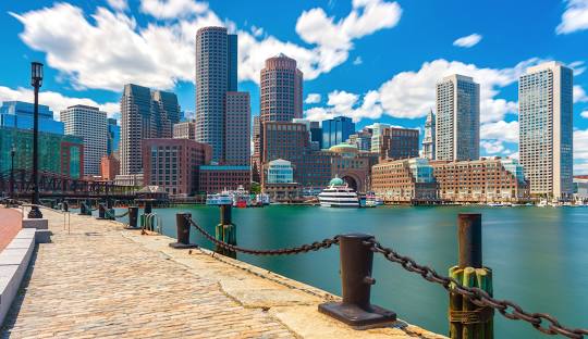 A view of Boston's waterfront with tall city buildings under a blue sky with scattered clouds, a cobblestone path, and a chain link fence along the water's edge, showcasing the city's blend of historic charm and modern innovation like managed IT services in Massachusetts.