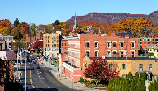 A small town with brick buildings, a tree-lined street, and colorful autumn foliage in the background, set against a backdrop of hills - where businesses thrive with managed IT services in Meriden, CT.