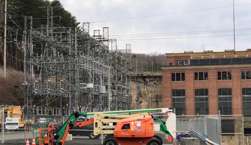 A utility site with metal structures and equipment near a brick building. An orange and white cherry picker is stationed in front of a wire fence, indicative of the area’s industrial activity. Managed IT services in Monroe CT ensure seamless operations. Trees and a hill are in the background.