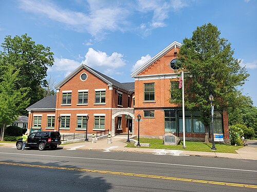 A two-story brick building with an arched entrance, adjacent lamp posts, a parked black SUV, and a visible flagpole, situated along a paved road on a sunny day in the heart of New Hartford CT.