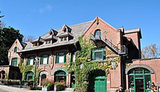 A three-story historic brick building with green-trimmed windows, dormers, and ivy growing up one side, set against a clear blue sky, stands as a testament to the charm of Norfolk CT.