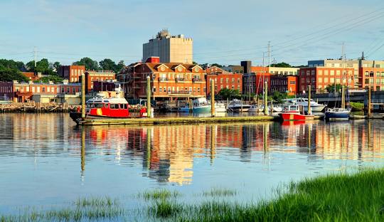 A waterfront scene in Norwalk, CT shows boats docked at a marina with buildings in the background, all reflected in the calm water. The area is known for its tranquility and also for its modern managed IT services.