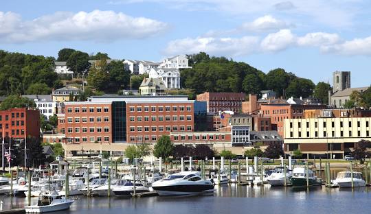 A waterfront marina with several boats docked, set against a backdrop of a small town with brown and yellow buildings and greenery on a hill under a blue sky with clouds, reflects the charming vibe of Norwich CT, where managed IT services ensure the tranquility remains undisturbed.