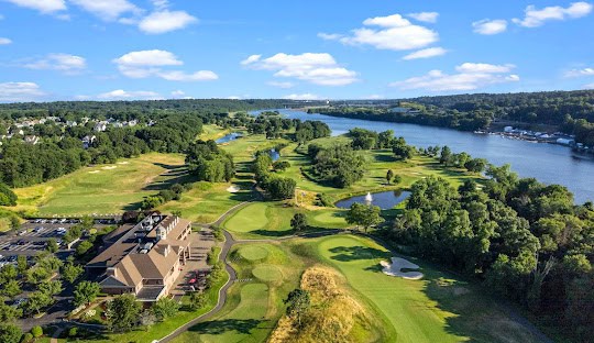 Aerial view of a sprawling golf course situated next to a river, with a large clubhouse on the left and a mix of green fairways, bunkers, and water features. Suburban homes are visible in the distance, epitomizing the serene charm that makes managed IT services in Orange CT an attractive option for businesses looking for tranquility.