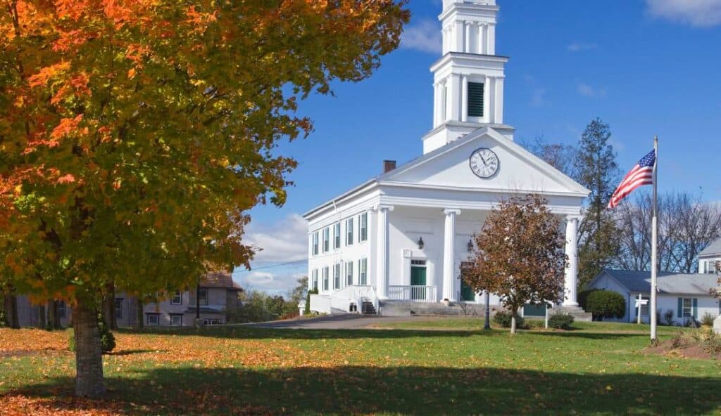 A white church with a clock tower is surrounded by autumn foliage in Plymouth, CT. The church is situated on a green lawn with an American flag to the right, exemplifying the quaint charm that managed IT services help preserve in this historic town.