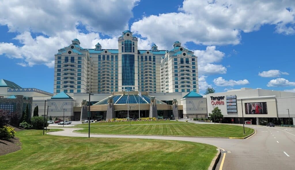 A large building with multiple floors and a glass pyramid structure at its center stands under a bright blue sky with clouds. Adjacent to it is a sign for “Tanger Outlets” and a video screen, showcasing the latest offerings from managed IT services in Preston, CT.