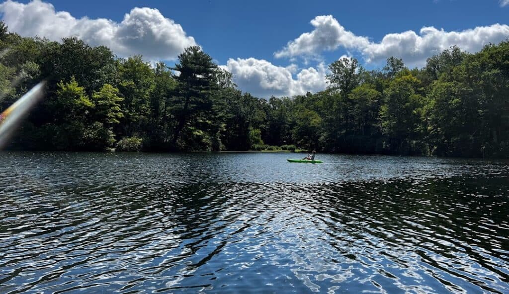 A person in a green kayak paddles on a calm, tree-lined lake under a partly cloudy sky, reminiscent of the serene landscapes around Redding CT.